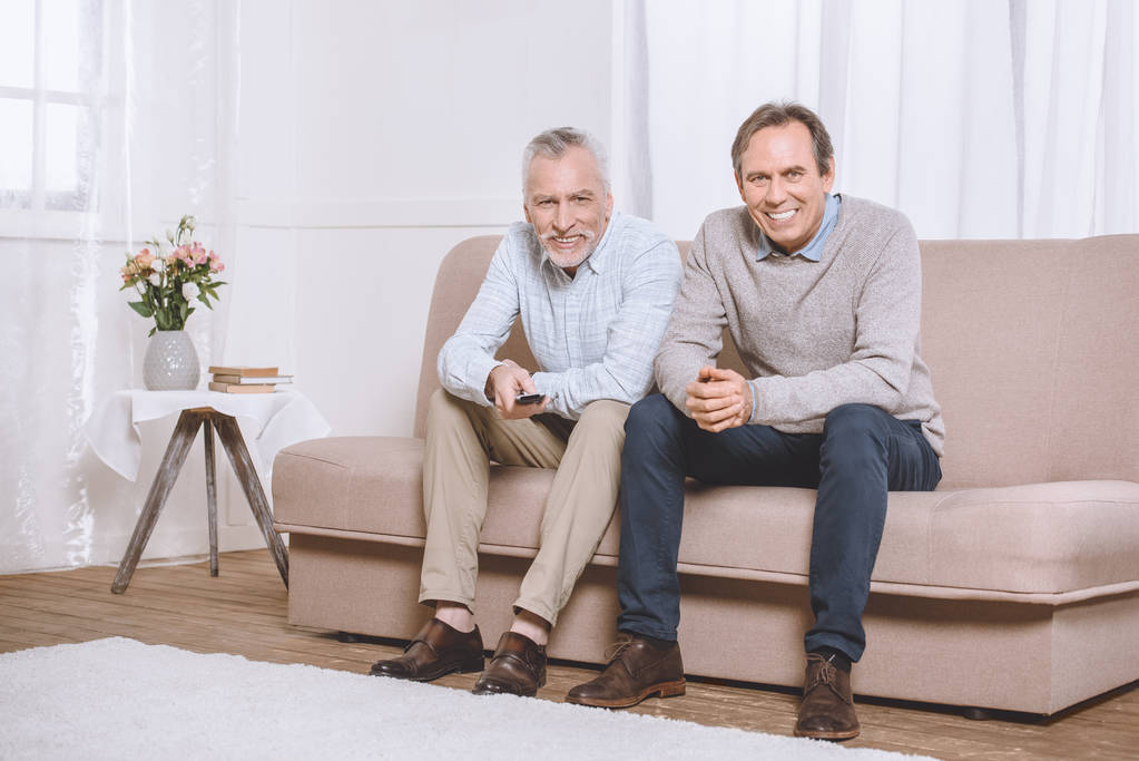 Two happy adult men sitting on sofa in light living room and watching tv - Photo, Image