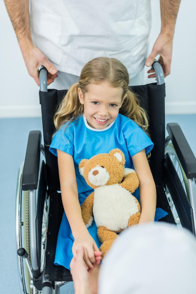 smiling kid sitting with soft toy on wheelchair and looking at camera - Photo, Image