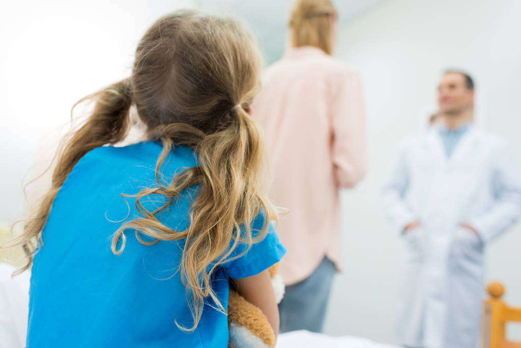 Rückansicht eines Kindes, das auf dem Krankenhausbett sitzt, während die Mutter mit dem Arzt spricht - Foto, Bild