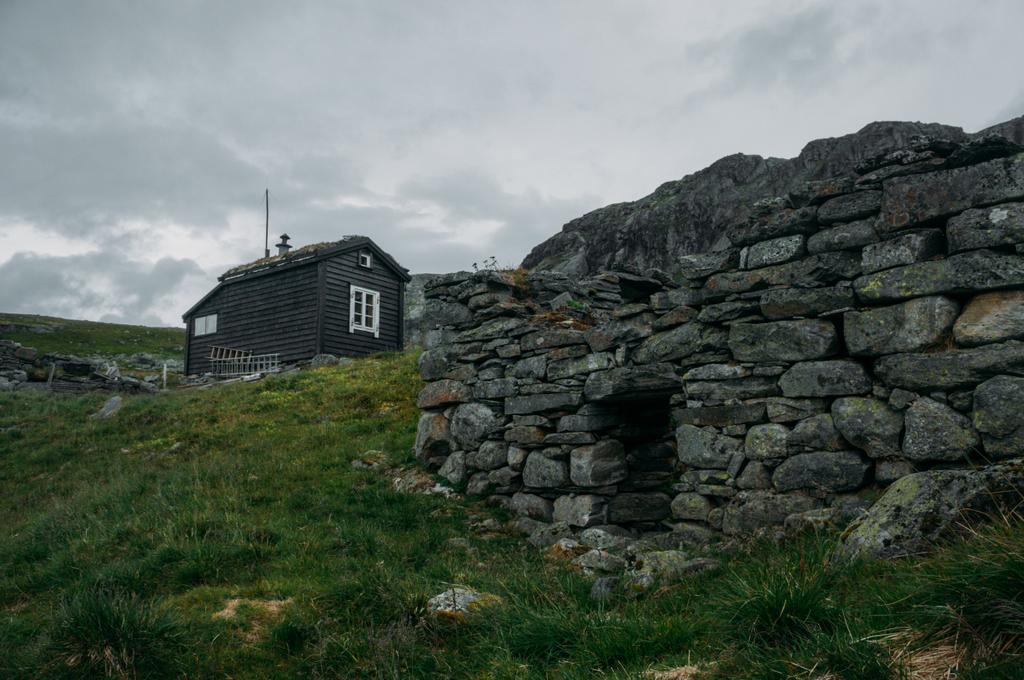 Ruinierte alte Steinmauer und ländliches Haus im Hintergrund über Feld mit grünem Gras, Norwegen, Hardangervidda Nationalpark - Foto, Bild