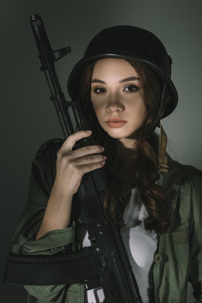 beautiful young woman in military helmet with rifle, on grey with shadows - Photo, Image