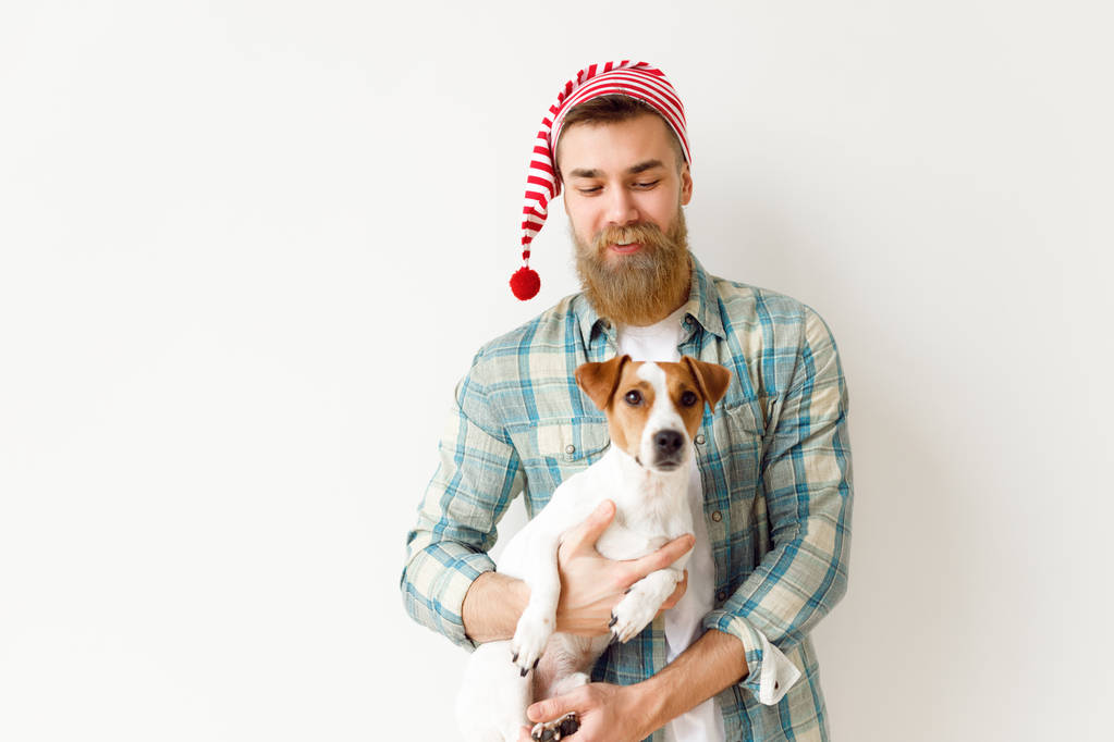 Позитивна бородата чоловіча модель носить святковий капелюх і картату сорочку, тримає свого улюбленого улюбленця, ізольованого на білому тлі. Собака як символ майбутнього Нового року. Свята та концепція святкування
 - Фото, зображення