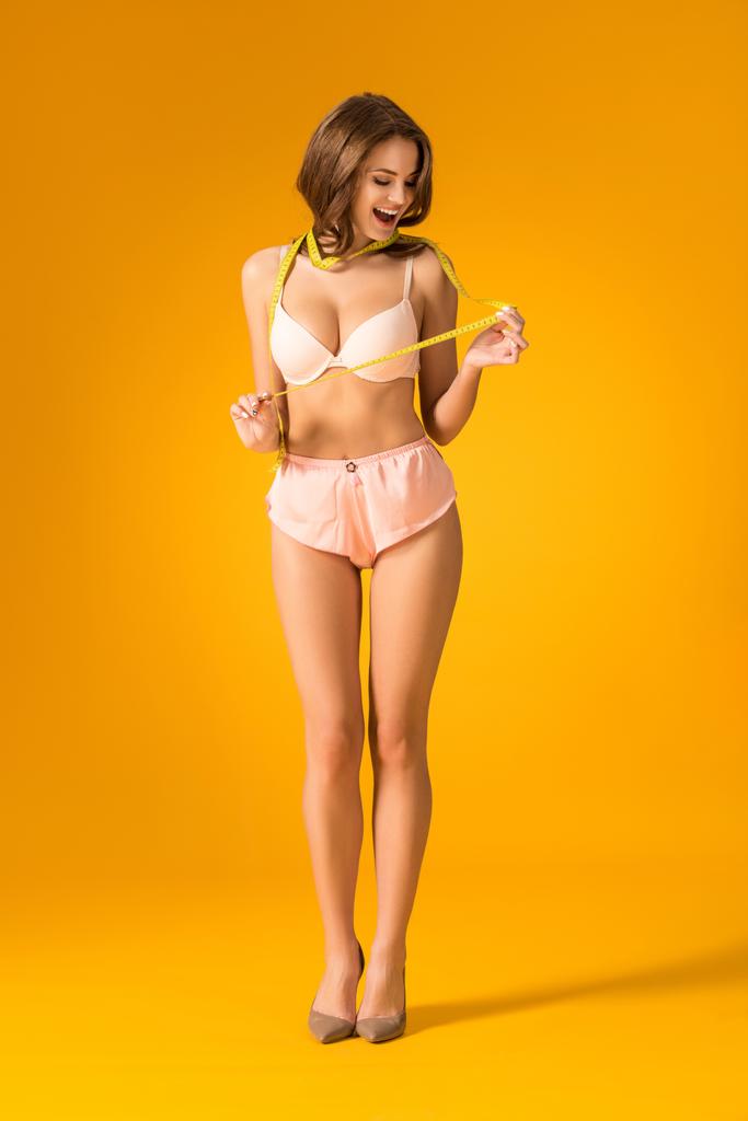 verbaasd sexy meisje in lingerie set meetlint kijken op oranje - Foto, afbeelding
