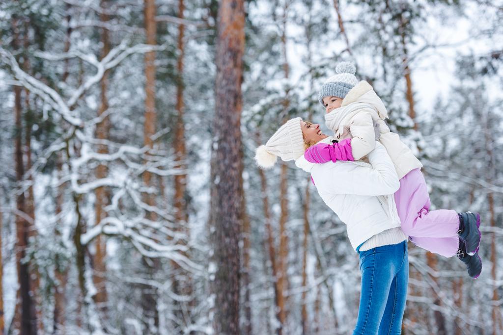 вид сбоку на мать, держащую маленькую дочь на руках в зимнем лесу
 - Фото, изображение