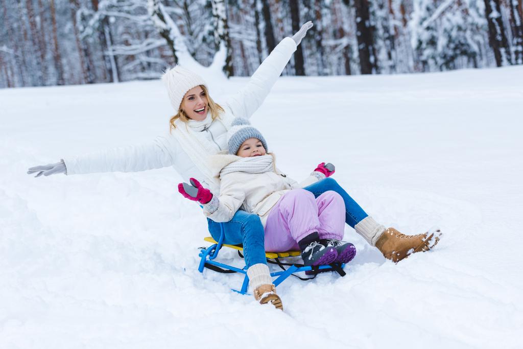 gelukkige dochter en moeder sleeën samen in winter park - Foto, afbeelding