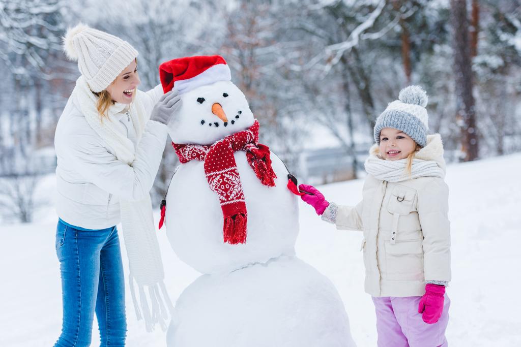 happy mother and daughter making snowman together in winter forest - Photo, Image