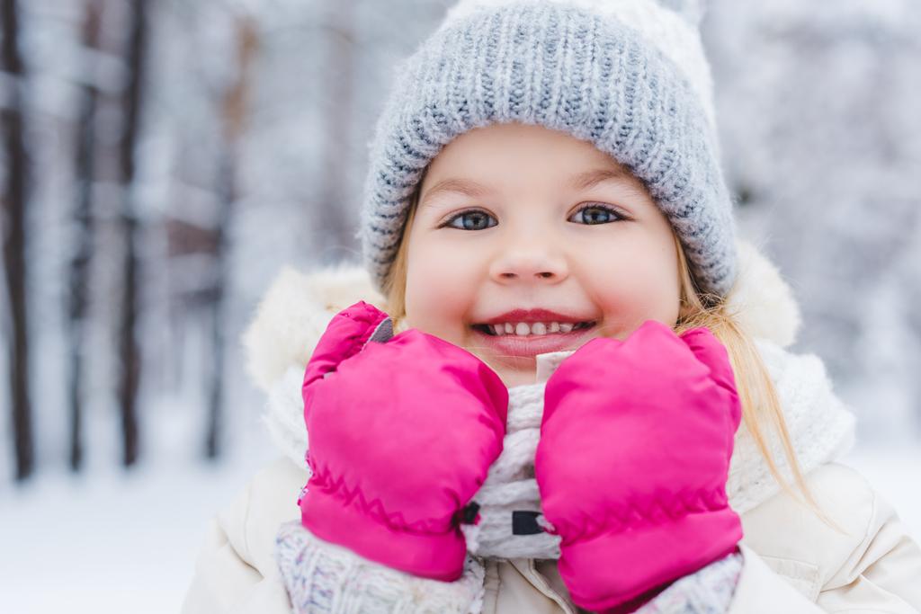 close-up portrait of cute little child in hat and mittens smiling at camera in winter park - Photo, Image