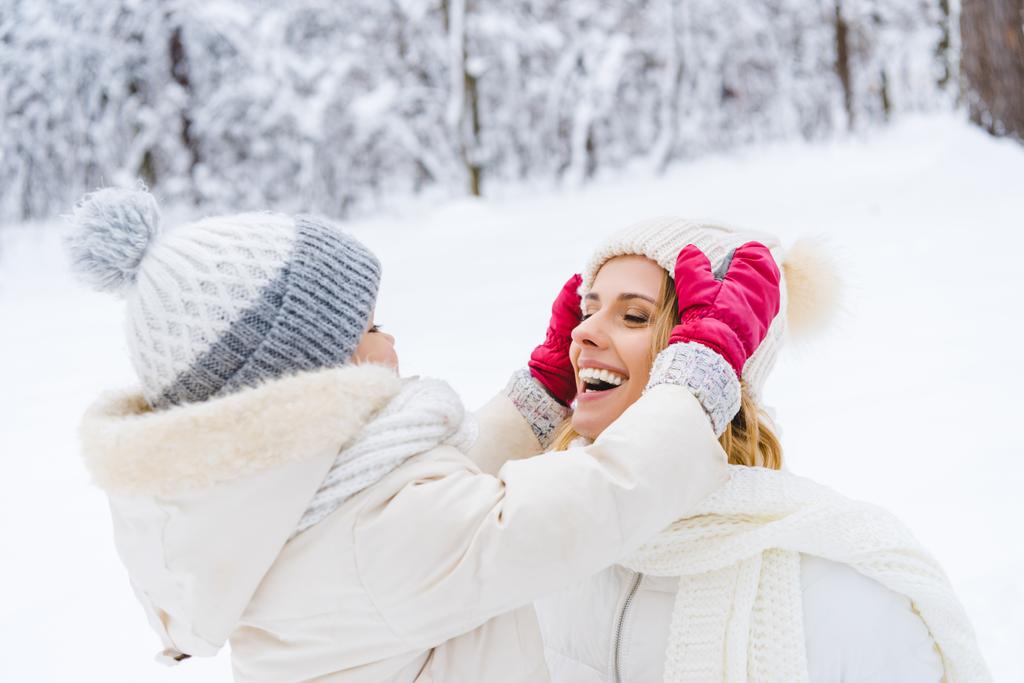 schöne glückliche Mutter und Tochter tragen Hüte und lächeln einander im Winterpark zu - Foto, Bild