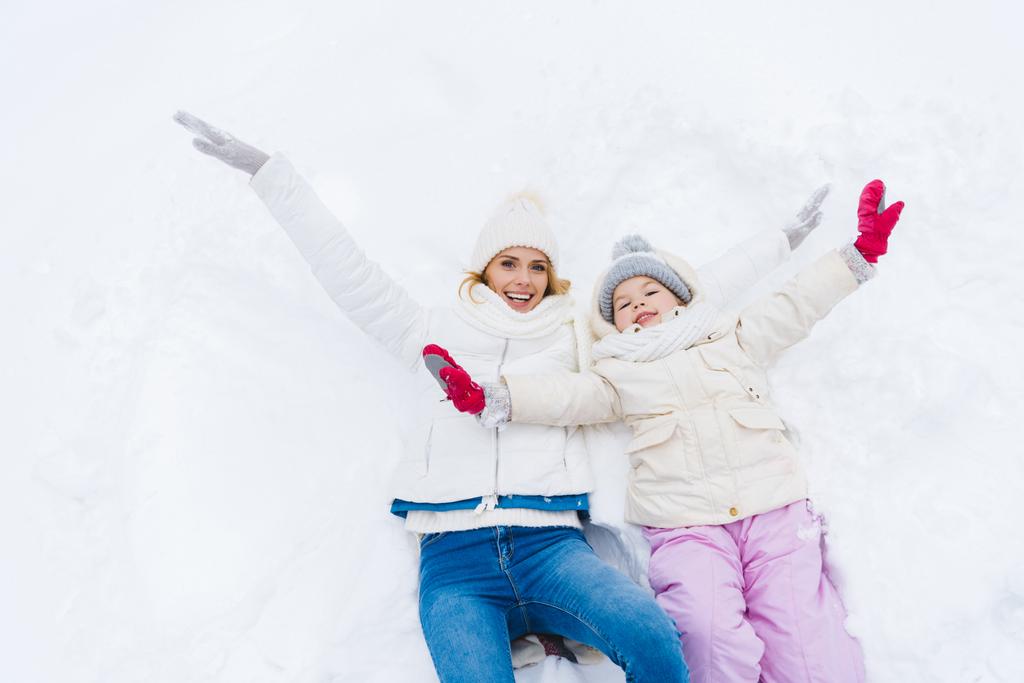 ευτυχισμένη μητέρα και κόρη με ανοιχτές αγκάλες χαμογελάει κάμερα ξαπλωμένοι μαζί στο χιόνι  - Φωτογραφία, εικόνα