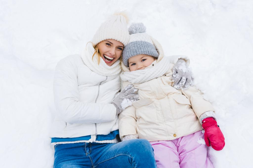 šťastný matka a dcera se usmívá na kameru při ležení společně ve sněhu  - Fotografie, Obrázek