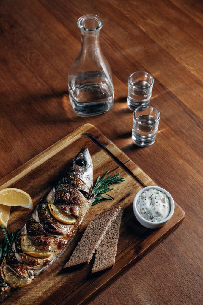 Close-up shot van gegrilde vis met citroen op een houten bord met wodka in karaf en shots - Foto, afbeelding