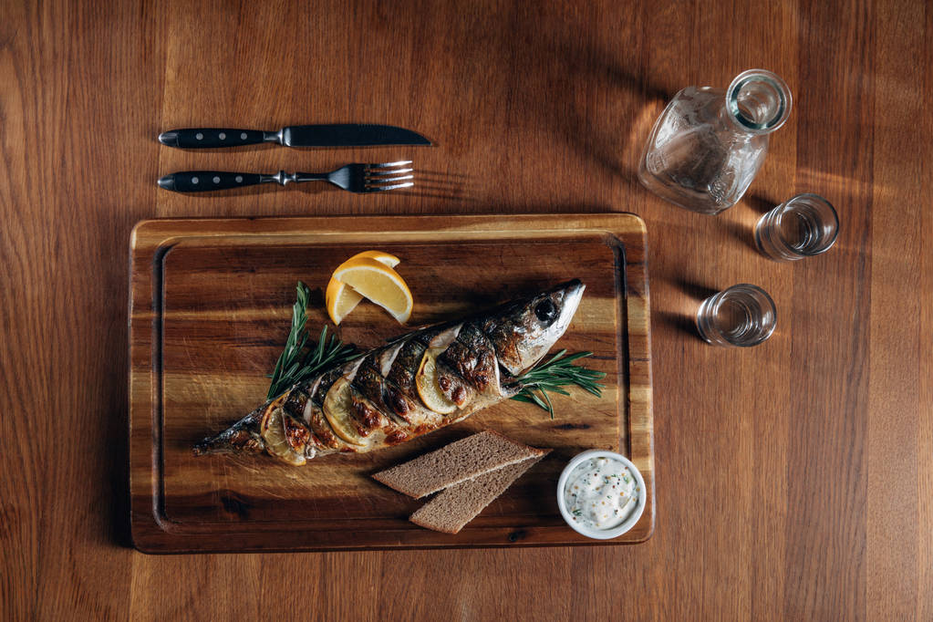 Draufsicht auf gegrillten Fisch mit Zitrone auf Holzbrett mit Wodka im Dekanter und Shots - Foto, Bild