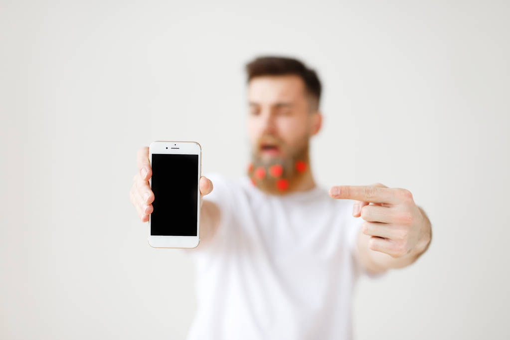 Moderno smartphone con schermo bianco pubblicizzato dal modello maschile barbuto alla moda. Dispositivo elettronico nelle mani dell'uomo. Concentrazione selettiva sul gadget. Persone, tecnologie innovative e concetto di pubblicità
 - Foto, immagini