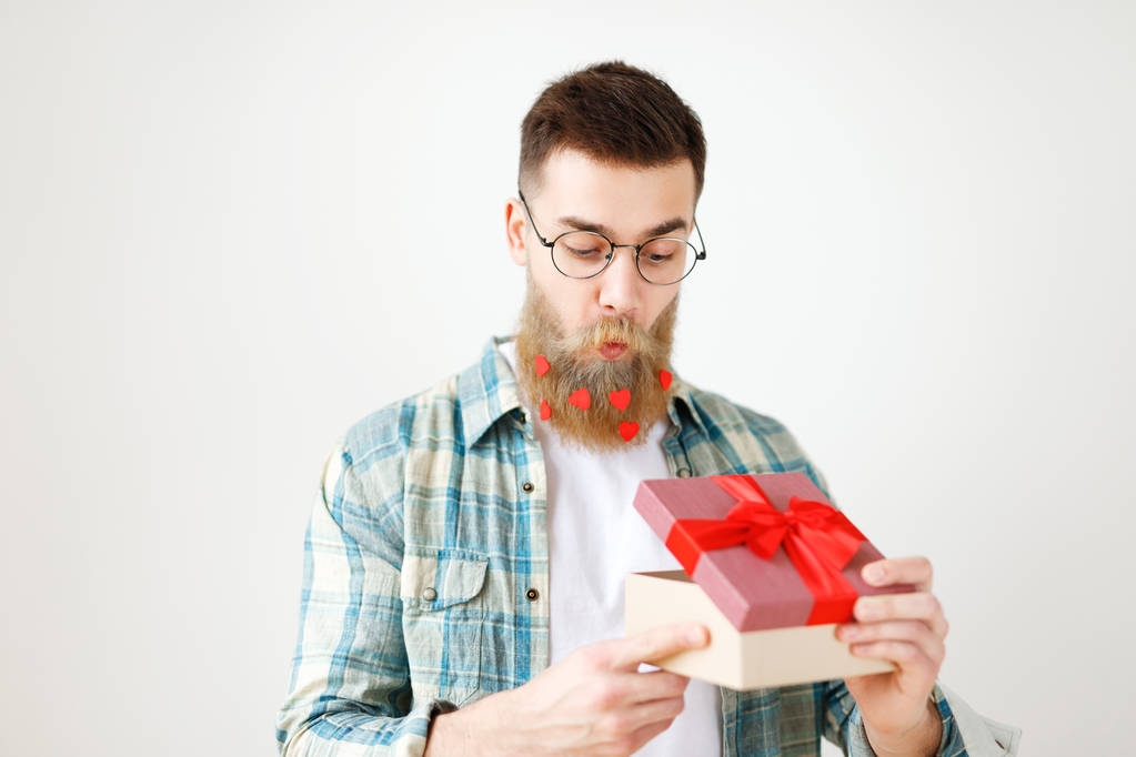 Hombre guapo sorprendido con barba gruesa y bigote mira con expresión intrigante como abre envuelto caja de regalo, siendo curioso lo que hay, lleva gafas redondas y camisa a cuadros, aislado
 - Foto, imagen