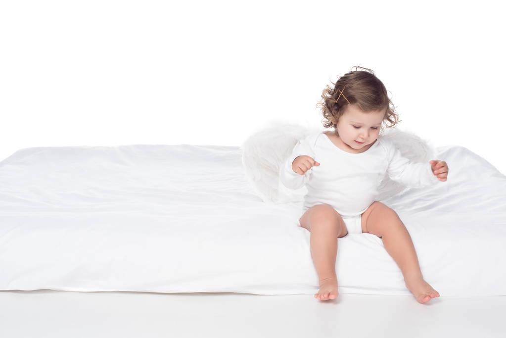 adorable happy baby with wings sitting on bed, isolated on white - Photo, Image