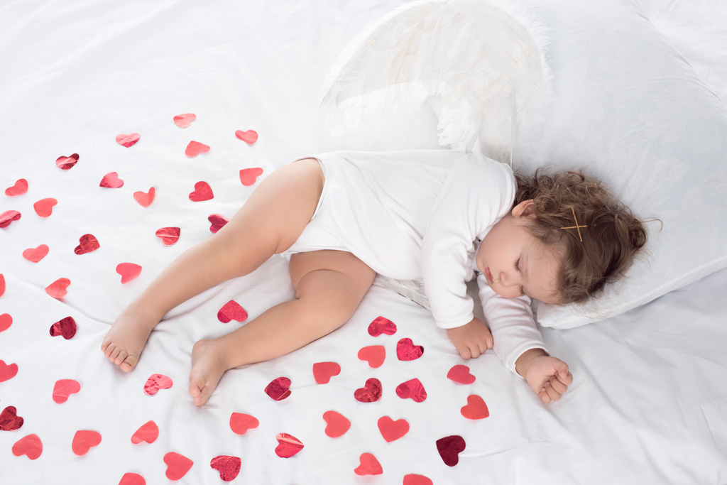 little cupid with wings sleeping on bed with red hearts - Photo, Image