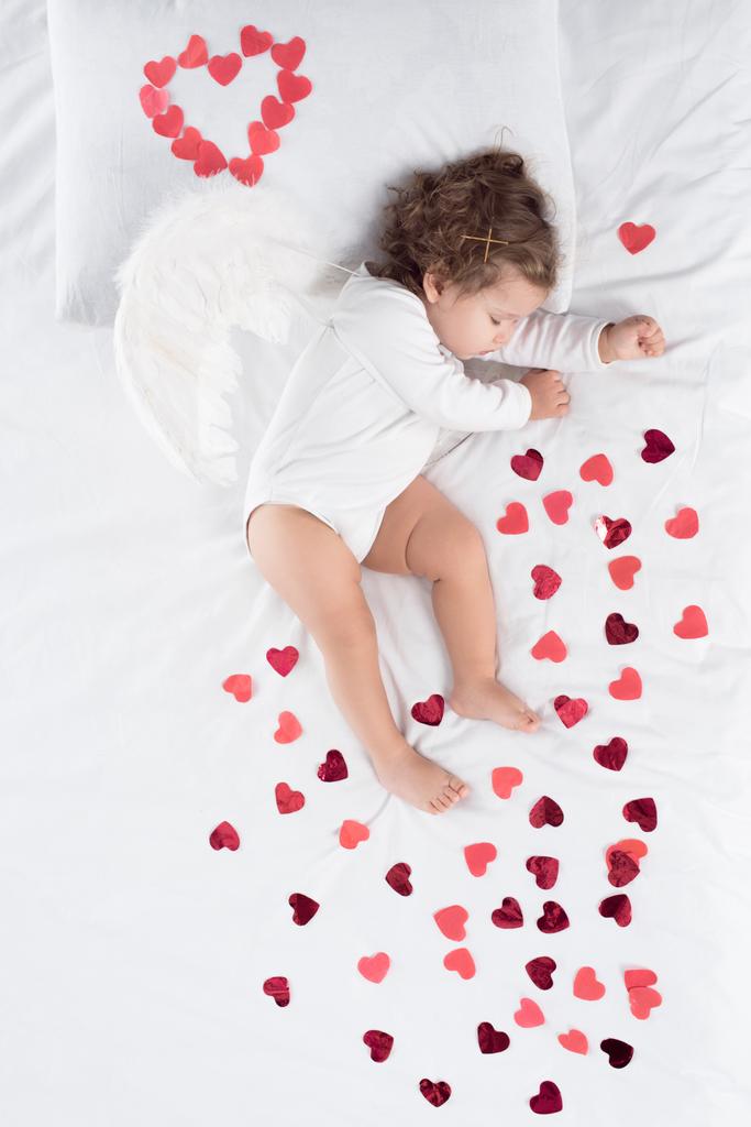 μικρό παιδί με φτερά στον ύπνο σε κρεβάτι με κόκκινες καρδιές - Φωτογραφία, εικόνα
