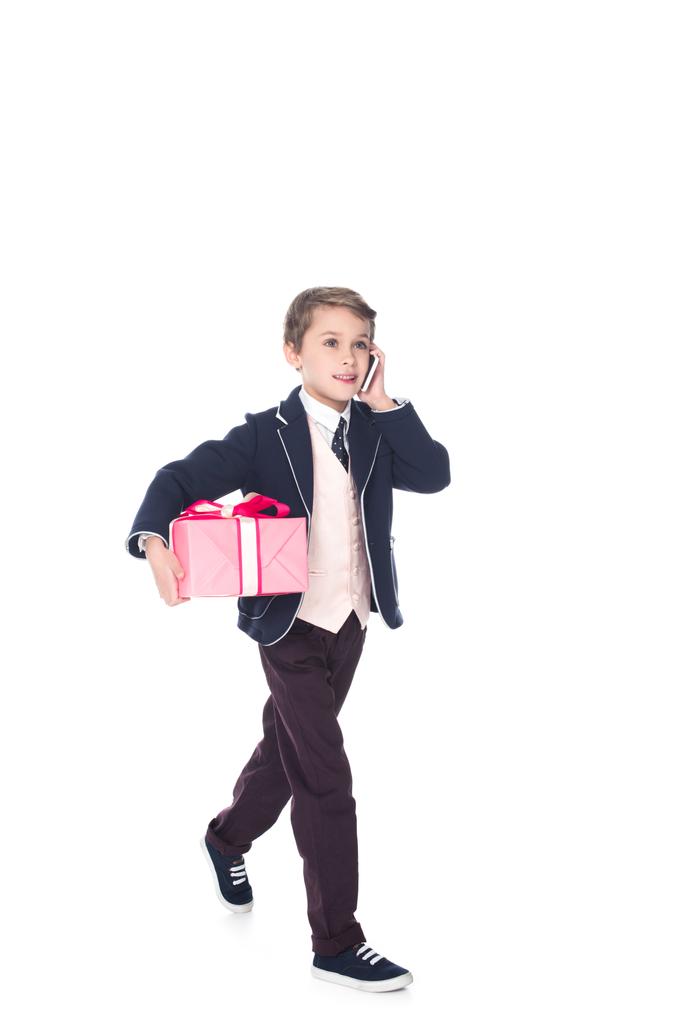 niño sonriente hablando en el teléfono inteligente y sosteniendo la caja de regalo mientras camina aislado en blanco
 - Foto, Imagen
