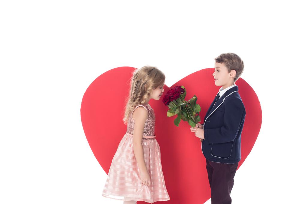 вид сбоку маленького мальчика, дарящего цветы красивой маленькой девочке и большой символ красного сердца сзади изолированный на белом
  - Фото, изображение