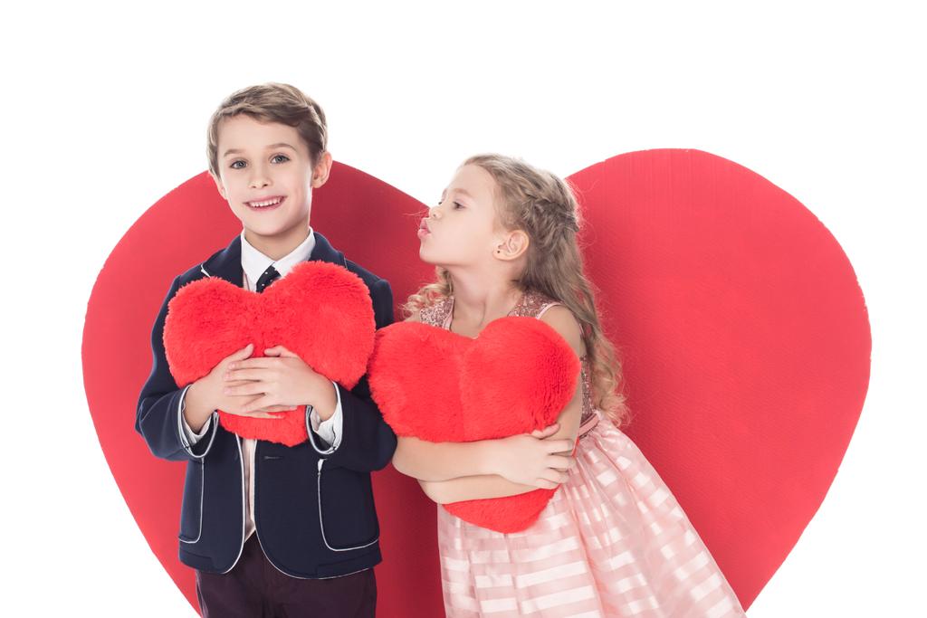 χαριτωμένα παιδάκια, κρατώντας την καρδιά σχήμα μαξιλάρια και σε θέση να φιλήσεις απομονωθεί σε λευκό  - Φωτογραφία, εικόνα