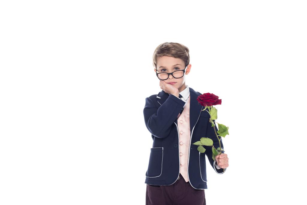 σοβαρά μικρό αγόρι σε κοστούμι και γυαλιά κρατώντας τριαντάφυλλο και βλέπουν τα φωτογραφικών μηχανών που απομονώνονται σε λευκό - Φωτογραφία, εικόνα