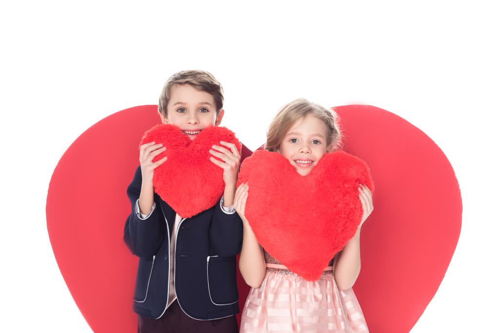 χαριτωμένα παιδάκια κρατώντας μαξιλάρια σε σχήμα καρδιάς και να χαμογελά στη φωτογραφική μηχανή που απομονώνονται σε λευκό  - Φωτογραφία, εικόνα