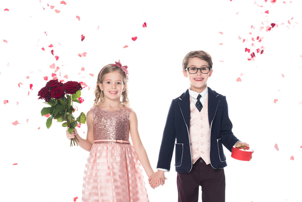 schöne stylische Kinder mit Rosen und herzförmigem Geschenkkarton, Händchen haltend und lächelnd in die Kamera auf weiß     - Foto, Bild