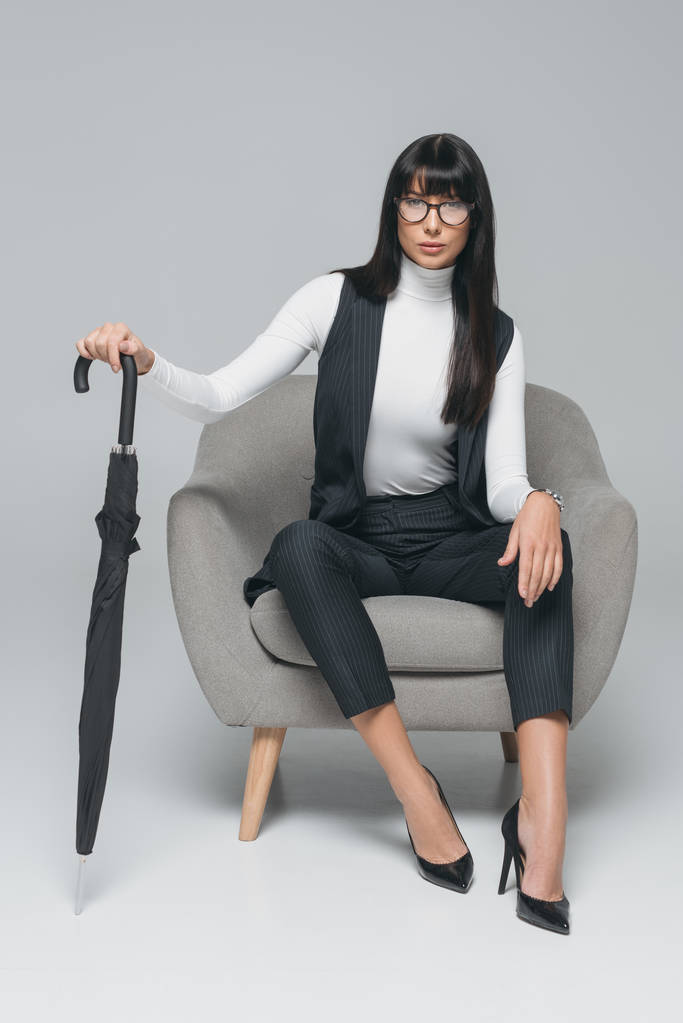 atraktivní bruneta podnikatelka sedí na křesle s deštníkem na šedé - Fotografie, Obrázek