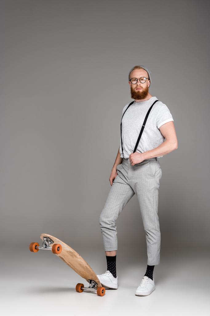hombre barbudo con estilo en tirantes de pie con monopatín y mirando a la cámara en gris
 - Foto, Imagen