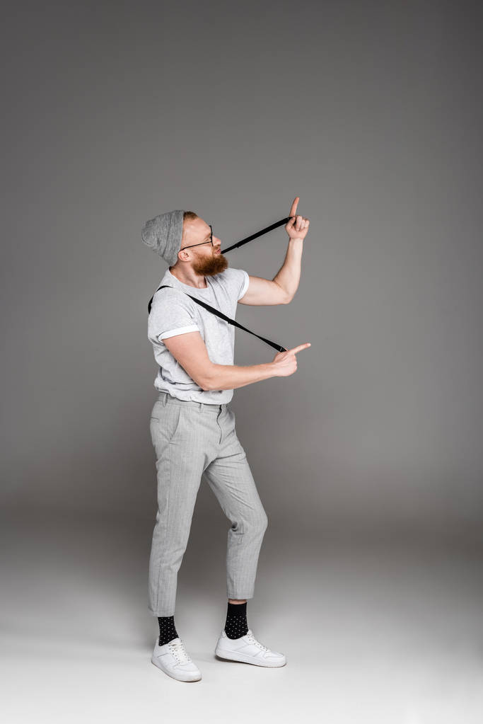 stijlvolle bebaarde man spelen met jarretels en wijzen met vingers op grijs  - Foto, afbeelding