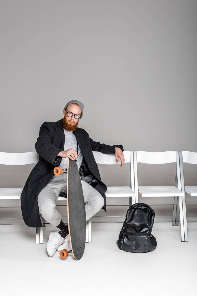 homme élégant en manteau et lunettes tenant skateboard tout en étant assis sur une chaise sur gris
 - Photo, image