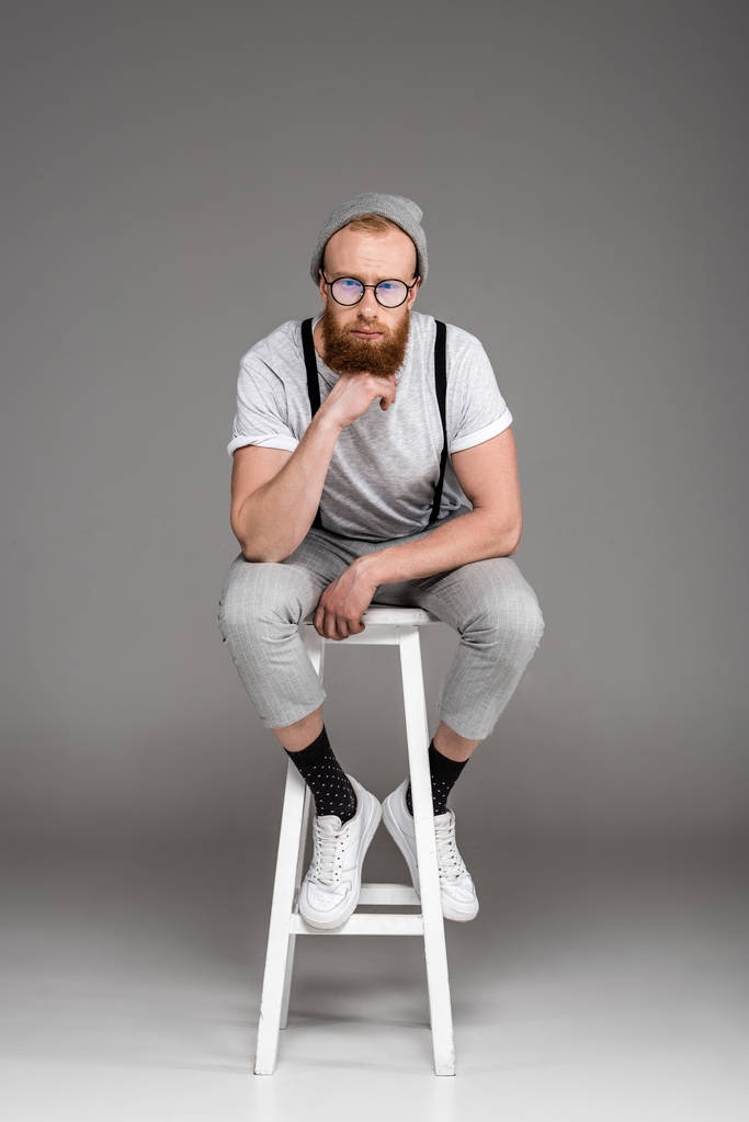stijlvolle bebaarde man in eyeglasees zit op kruk en camera op grijs   - Foto, afbeelding