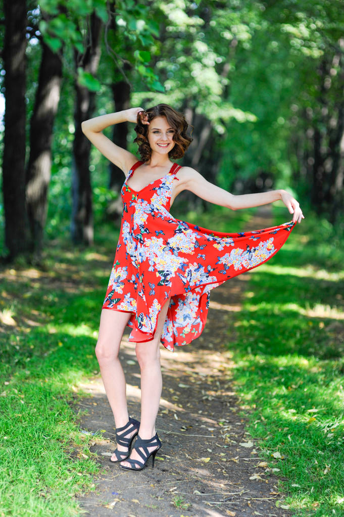 Een jonge brunette meisje in een rode jurk is dansen op een weg in een zomer-park tegen een achtergrond van bomen - Foto, afbeelding