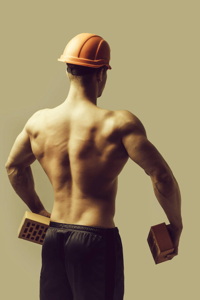 セクシーな筋肉運動の強力なボディを持つ若いハンサムなマッチョな男のビルダーが裸の胴体と強力なバック勝利オレンジ ハード帽子またはヘルメットは、灰色の背景のレンガを保持しています。 - 写真・画像