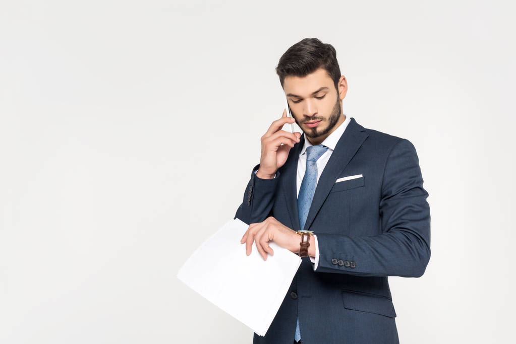 joven hombre de negocios sosteniendo papeles y hablando en el teléfono inteligente mientras comprueba reloj de pulsera aislado en gris
 - Foto, imagen