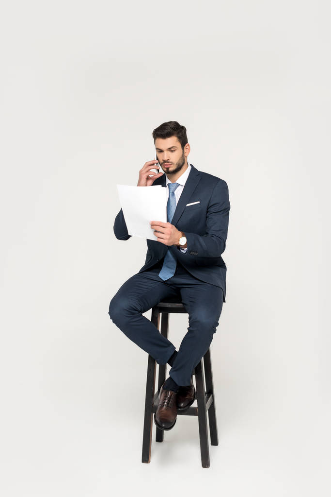 серьезный молодой бизнесмен разговаривает на смартфоне и читает газеты, сидя на стуле изолированы на серый
 - Фото, изображение