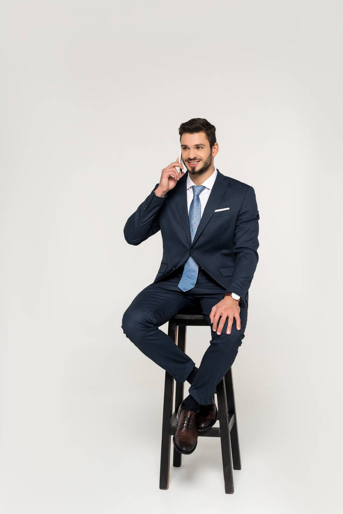 lachende jonge zakenman praten op smartphone terwijl zittend op een kruk en op zoek weg geïsoleerd op grijs - Foto, afbeelding