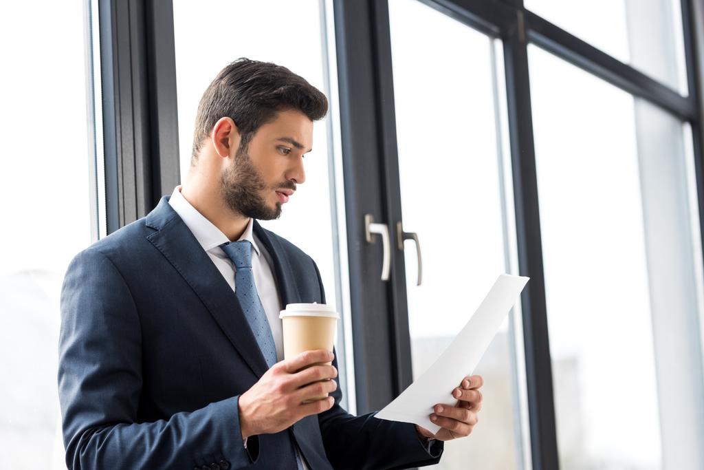 młody biznesmen posiadający kawa na wynos i czytanie dokumentów w biurze - Zdjęcie, obraz