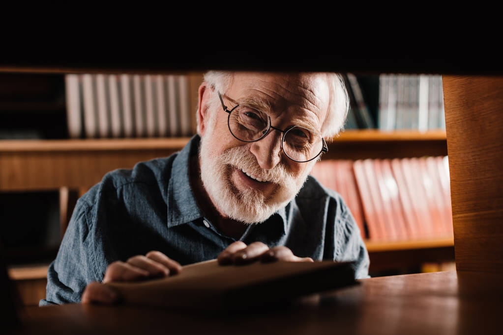 hymyilevä harmaa tukka kirjastonhoitaja katselee kameraa hyllyn läpi
 - Valokuva, kuva