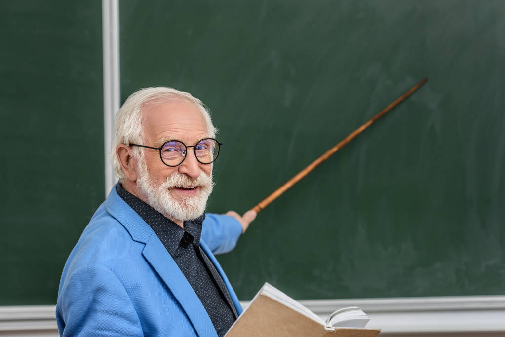 Седой профессор по волосам держит книгу и указывает на что-то на доске
  - Фото, изображение