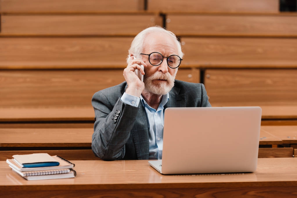 siwe włosy profesor rozmawia przez smartfona i patrząc na laptopa - Zdjęcie, obraz
