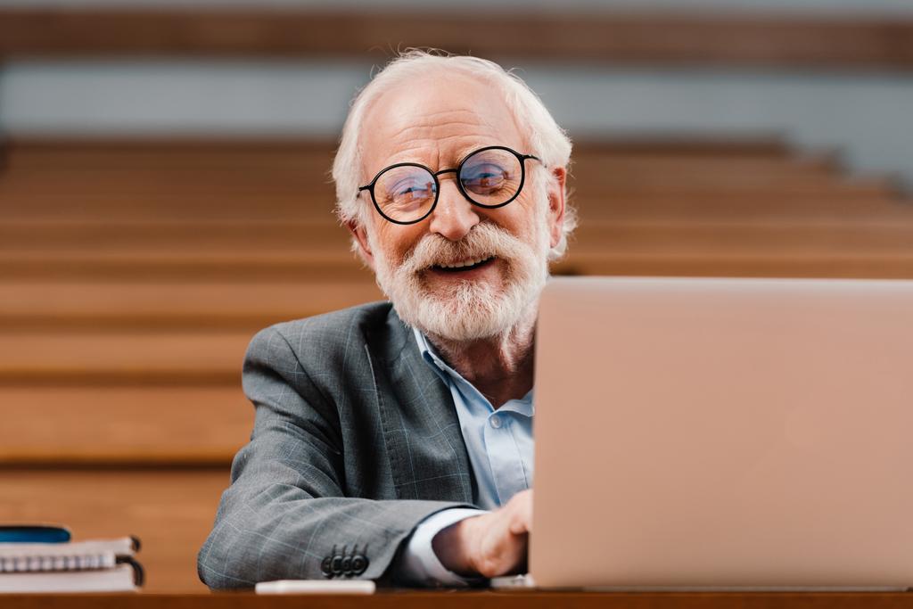 usmívající se šedými vlasy profesor sedí v prázdné místnosti s notebookem - Fotografie, Obrázek
