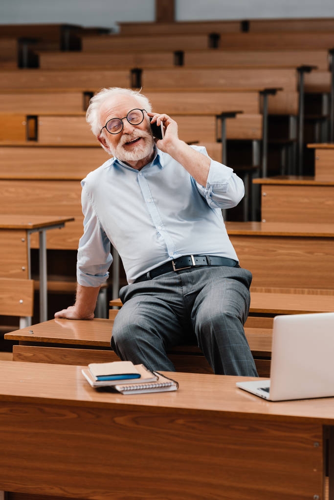 usmívající se šedými vlasy profesor sedí na stole v prázdné místnosti a mluví o smartphone - Fotografie, Obrázek