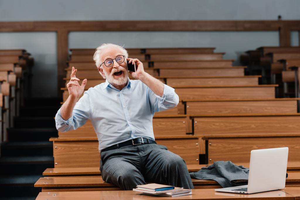 professeur de cheveux gris souriant assis sur le bureau dans la salle de conférence vide et parlant par smartphone
 - Photo, image