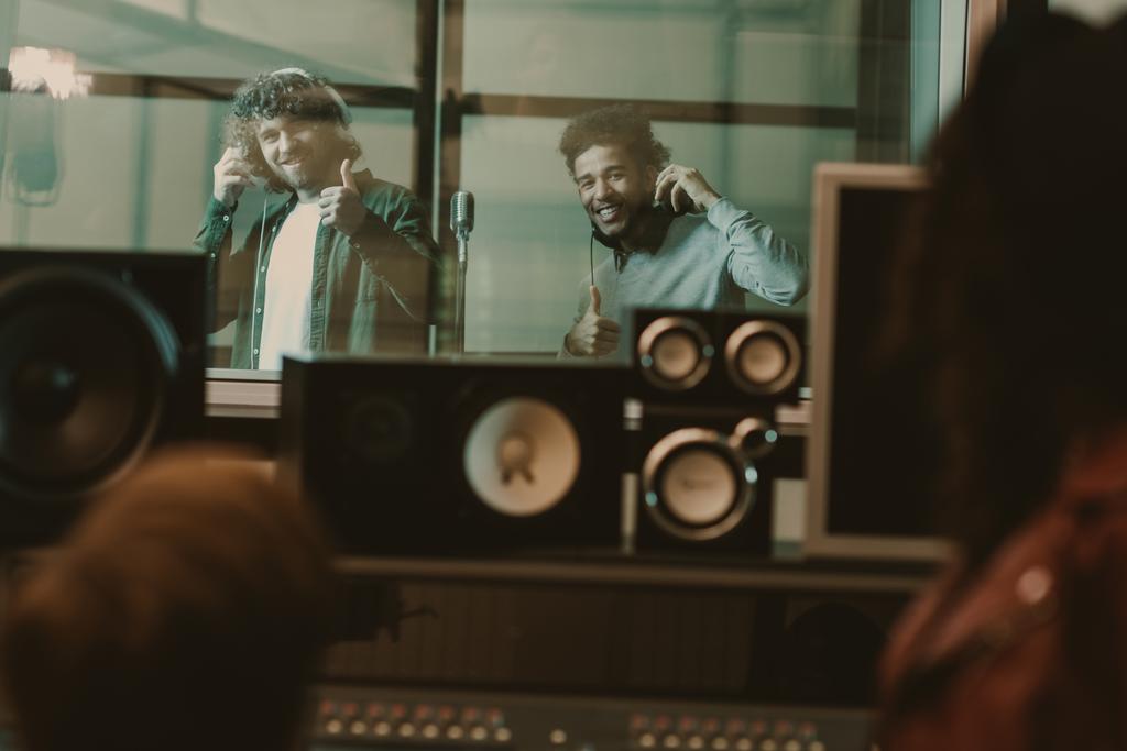 young handsome musicians recording song behind glass at studio and showing thumbs up - Photo, Image