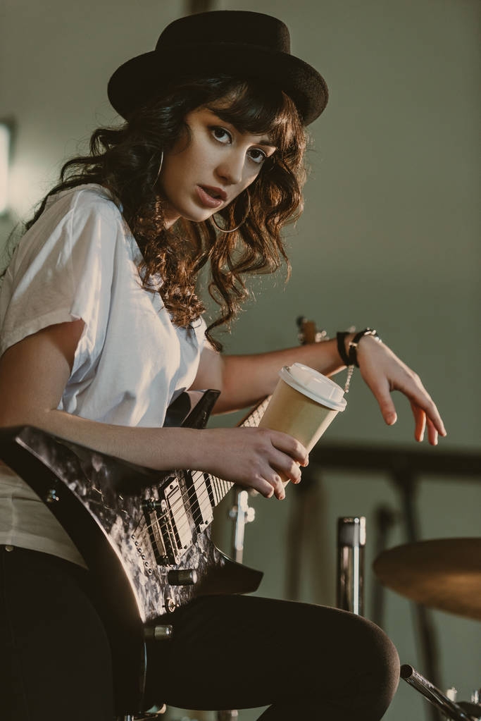 beautiful female musician with electric guitar and cup of coffee to go looking at camera - Photo, Image
