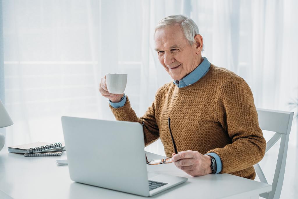 Старший уверенный в себе человек работает на ноутбуке и держит чашку кофе
 - Фото, изображение