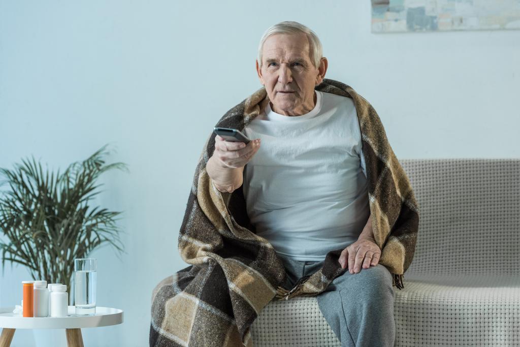 Άρρωστος ανώτερος άνθρωπος που καλύπτονται σε καρό ρολόγια τηλεόραση στο δωμάτιο με τα φάρμακα στον πίνακα - Φωτογραφία, εικόνα