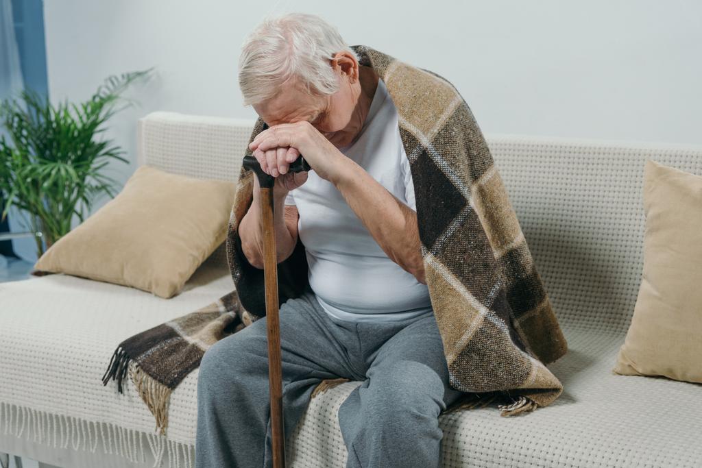 Väsynyt vanhempi mies yllään ruudullinen nojaa keppi istuessaan sohvalla
 - Valokuva, kuva