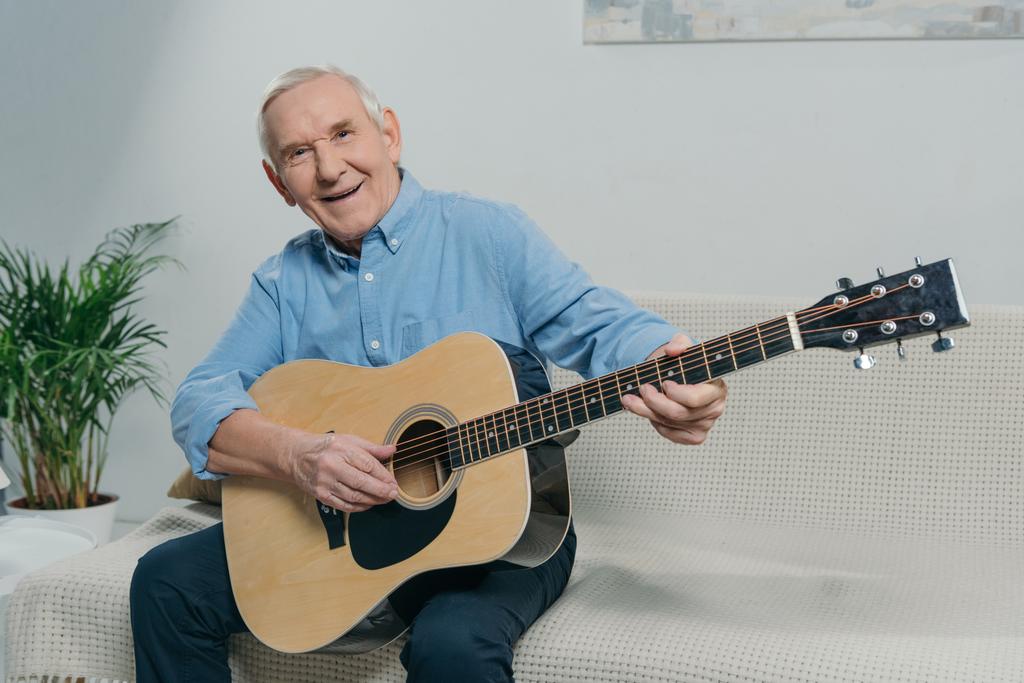 Старший счастливый человек играет на акустической гитаре, сидя на диване в комнате
 - Фото, изображение